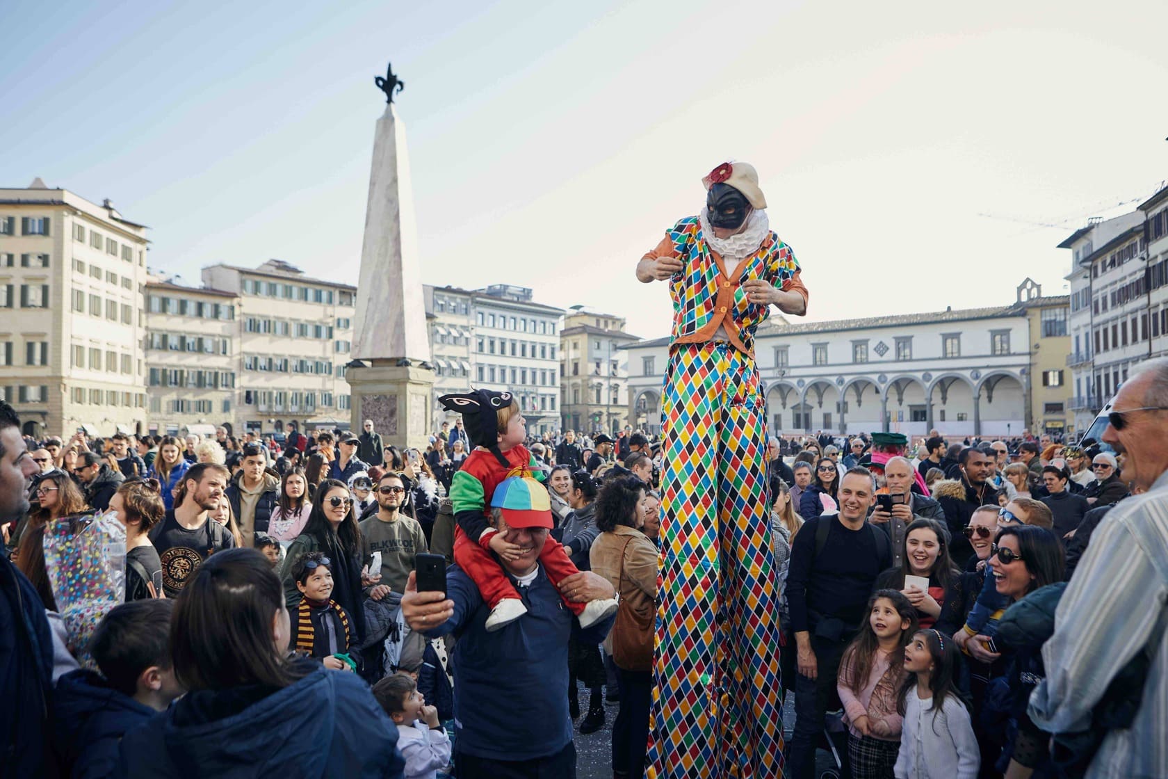 Carnevale in una piazza a Firenze: Clown su trampoli in costume da arlecchino che diverte la folla.
